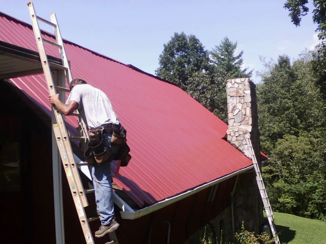 Roofer on a ladder beside a red metal roof and stone chimney — Soffit & Fascia Repair by Roofing Brothers in Alsea